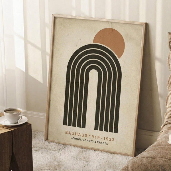 May include: Framed vintage-style poster leaning against white wall in cozy room setting. Design features cream background with dark green concentric arched lines forming rainbow shape, topped by rust orange circle resembling sun. Text reads "Bauhaus 1919-1933 School of Arts & Crafts" in brown lettering. Light wood frame, white coffee cup on wooden stool nearby, soft beige blanket visible, natural window light creating shadows.
