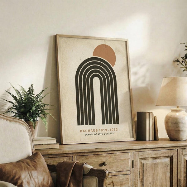 May include: Framed minimalist art print featuring black concentric arches with terracotta circle above, displayed on rustic wooden sideboard. Room scene shows neutral walls, fern plant in white pot, ceramic vase, stacked books, and cream upholstered chair with brown leather cushions. Natural lighting creates warm, cozy atmosphere in modern farmhouse style interior.