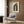 May include: Framed Bauhaus poster with geometric rainbow arch and orange sun design mounted on wood-paneled wall above beige upholstered armchair with patterned throw pillow, brass floor lamp, and dark round side table with open book in modern living room setting