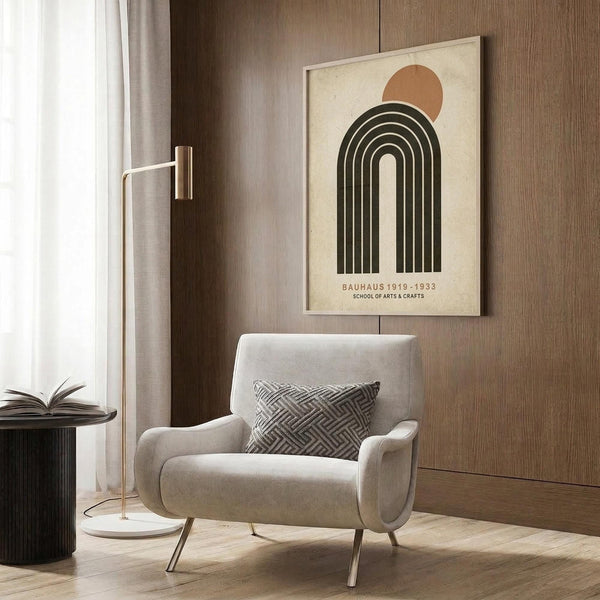 May include: Framed Bauhaus poster with geometric rainbow arch and orange sun design mounted on wood-paneled wall above beige upholstered armchair with patterned throw pillow, brass floor lamp, and dark round side table with open book in modern living room setting