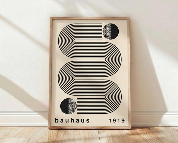 May include: Framed geometric poster with black striped wavy lines forming an S-shaped pattern on cream background, featuring two black circles with concentric line details, displayed against white textured wall on light wood floor with natural window light shadows
