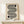 May include: Framed Bauhaus-style geometric poster with black flowing wavy lines and two half-circles on cream background, displayed on light wood console table beside white wrapped gift with twine and small greenery sprig, against neutral wall with natural lighting