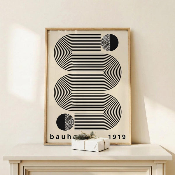 May include: Framed Bauhaus-style geometric poster with black flowing wavy lines and two half-circles on cream background, displayed on light wood console table beside white wrapped gift with twine and small greenery sprig, against neutral wall with natural lighting