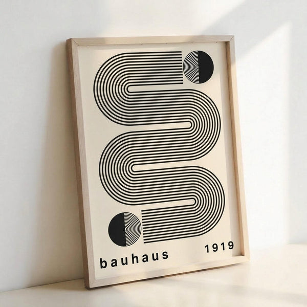 May include: Framed geometric poster with black and cream design featuring flowing curved lines forming an abstract serpentine pattern, two half-circles with concentric rings at top and bottom corners, text reading "bauhaus 1919" in lowercase letters, displayed in light wooden frame leaning against white wall with soft lighting casting shadows