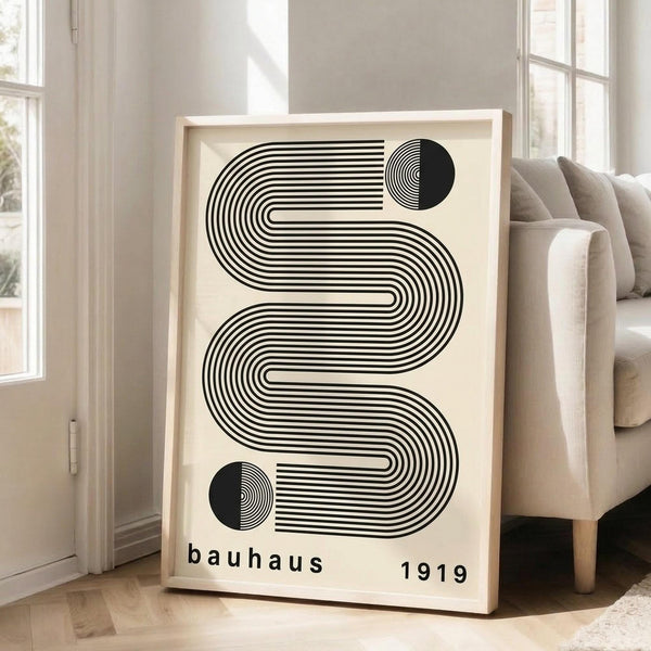 May include: Large framed print with black and white geometric design featuring flowing curved lines and circular elements on cream background, displayed on wooden floor against white wall next to beige upholstered sofa in bright living room with natural light from windows