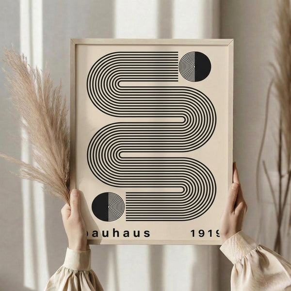 May include: Person in beige sweater holding framed geometric art print with black wavy lines and circles on cream background. Light wood frame, dried pampas grass in hand, soft natural lighting with white curtains in background. Modern minimalist Bauhaus-inspired design with flowing striped pattern.