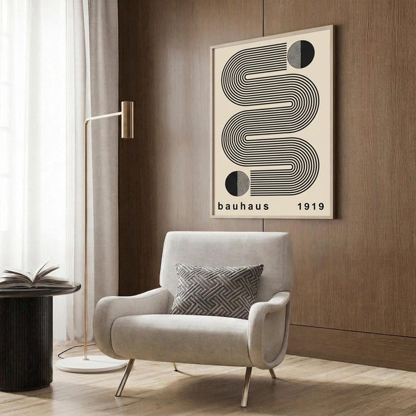 May include: A cream-colored poster with black geometric wavy lines and circular elements, text reading "bauhaus 1919" displayed in a wood-paneled room setting above a beige upholstered armchair with geometric patterned pillow, brass floor lamp, and dark round side table with open book