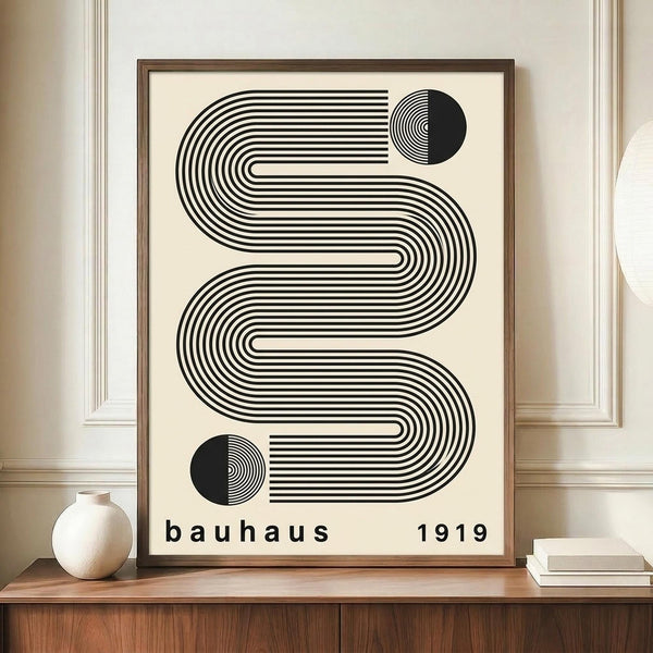 May include: Black and white geometric poster with flowing curved lines forming an S-like shape and two half-circles, text reading "bauhaus 1919" below, displayed in brown wooden frame on wooden sideboard next to white ceramic vase and books in cream-walled room with white trim molding.