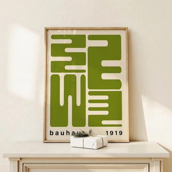 May include: Framed geometric poster with olive green abstract shapes forming interlocking patterns on cream background, displayed on light wood console table against white wall. Small wrapped gift box with evergreen sprig sits nearby. Design features bold angular forms and negative space in mid-century modern style.
