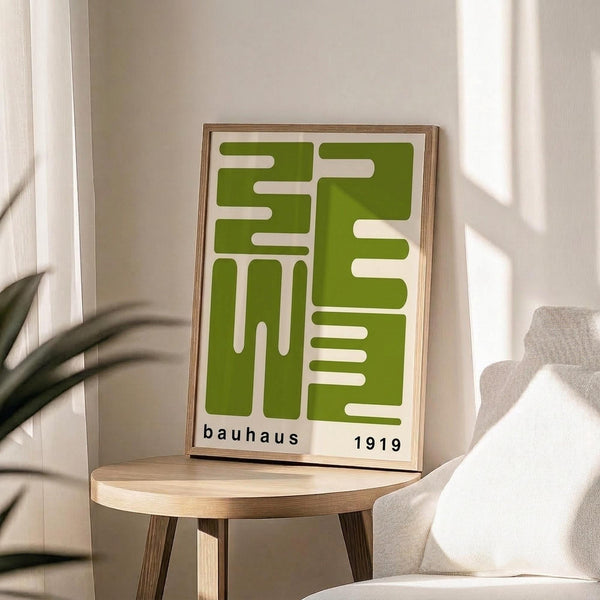 May include: Green geometric abstract poster with white interlocking shapes in wooden frame, displayed on round wooden stool next to beige upholstered chair. Sunlight casts shadows across neutral-toned room with plant visible in corner. The artwork features bold modernist design with "bauhaus 1919" text at bottom.
