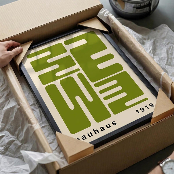 May include: Green and cream geometric Bauhaus poster in black frame being unpacked from cardboard box with tissue paper, showing bold angular shapes and "1919" text, displayed on white surface with wooden rolling pin nearby