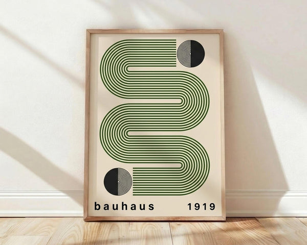 May include: Framed poster with green wavy striped design and black circular elements on cream background, displayed against white textured wall on light wood floor. Natural lighting creates soft shadows across the scene. The artwork features flowing geometric lines that create an S-shaped pattern with "bauhaus 1919" text at bottom in black lettering.