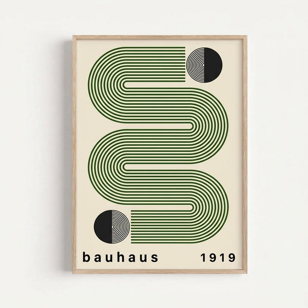 Framed geometric art print featuring flowing green striped waves in an S-curve pattern with two black semicircles containing concentric line details. The minimalist design is printed on cream paper with "bauhaus 1919" text below, displayed in a light wood frame against a white wall.