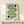 May include: Framed geometric poster with green wavy lines and black circular elements on cream background, displayed on light wood surface next to small wrapped gift with greenery, leaning against white wall in minimalist room setting.
