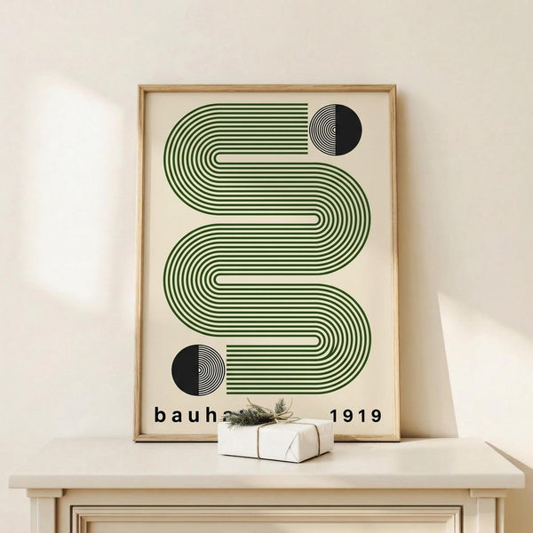 May include: Framed geometric poster with green wavy lines and black circular elements on cream background, displayed on light wood surface next to small wrapped gift with greenery, leaning against white wall in minimalist room setting.
