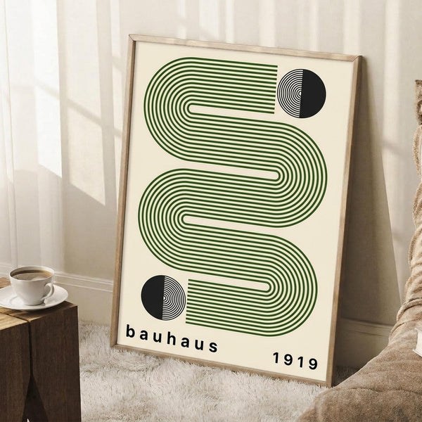 May include: A framed geometric poster with green flowing wave-like lines and black circular elements on cream background, displayed in a bright room corner next to a wooden side table with white coffee cup and saucer, leaning against white paneled walls on a textured rug.