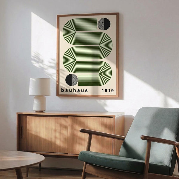 May include: Framed poster with green wavy parallel lines forming an S-shape and two black semicircles, displayed above a wooden sideboard with white ceramic lamp. Mid-century modern living room setting features sage green upholstered chair with wooden arms, round coffee table, and natural sunlight casting shadows on white wall.