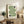 May include: Framed geometric art print with green wavy lines and black circles on cream background, displayed on rustic wooden sideboard beside fern plant, books, ceramic vase, and beige upholstered chair in neutral-toned living room setting.