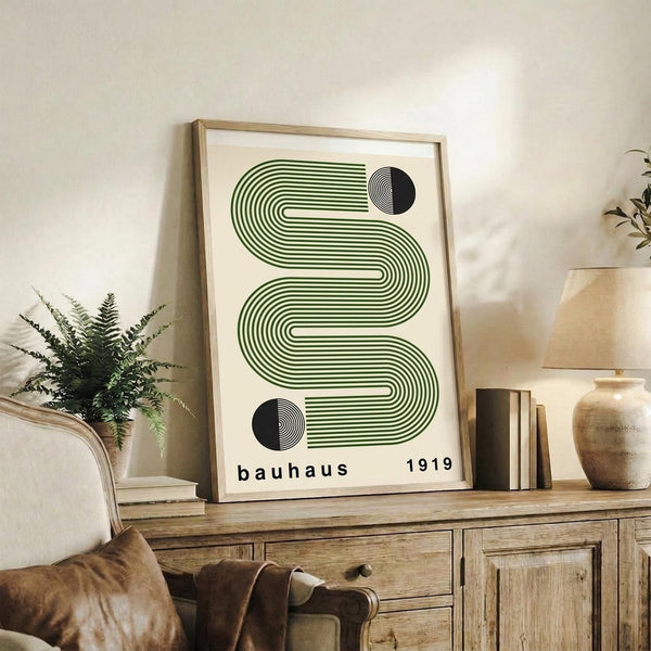 May include: Framed geometric art print with green wavy lines and black circles on cream background, displayed on rustic wooden sideboard beside fern plant, books, ceramic vase, and beige upholstered chair in neutral-toned living room setting.
