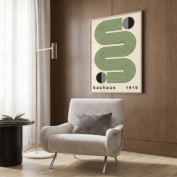 May include: Framed geometric poster with green wavy lines and black circular elements on cream background, displayed above beige upholstered chair with patterned pillow in modern living room setting with wood paneled walls, gold floor lamp, and dark round side table with open book.