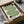 May include: Framed geometric poster with green wavy lines forming abstract shapes and black circular elements on cream background, displayed in black frame inside cardboard shipping box with protective wrapping and wooden dowels, styled on white fabric surface with glass vessel in background