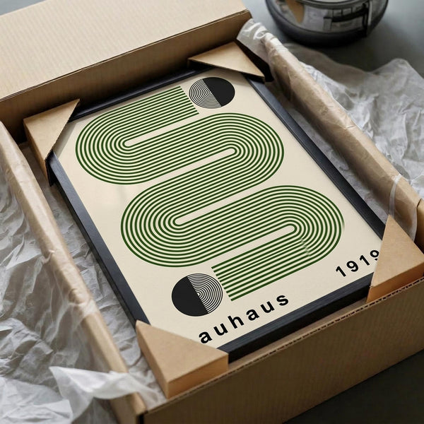 May include: Framed geometric poster with green wavy lines forming abstract shapes and black circular elements on cream background, displayed in black frame inside cardboard shipping box with protective wrapping and wooden dowels, styled on white fabric surface with glass vessel in background