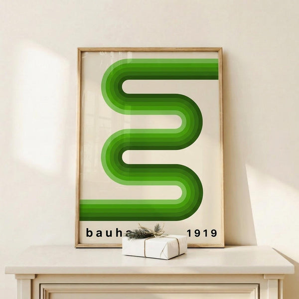 May include: Framed poster with green curved geometric shape resembling flowing ribbons or an S-curve against cream background, displaying "bauhaus 1919" text below. Natural wood frame sits on light wooden surface with small white wrapped gift box decorated with twine and greenery sprig. Neutral wall background creates minimalist styling setup.