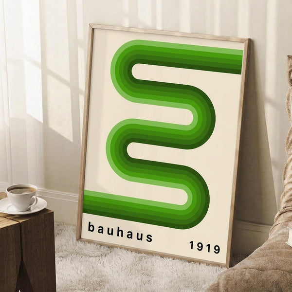 May include: Framed poster with green serpentine geometric design featuring flowing S-shaped curves with dimensional shading effects on cream background. Text reads "bauhaus 1919" in black lowercase letters. Light wood frame leans against white wall corner next to wooden side table with white coffee cup and saucer, beige textured throw visible at bottom right.