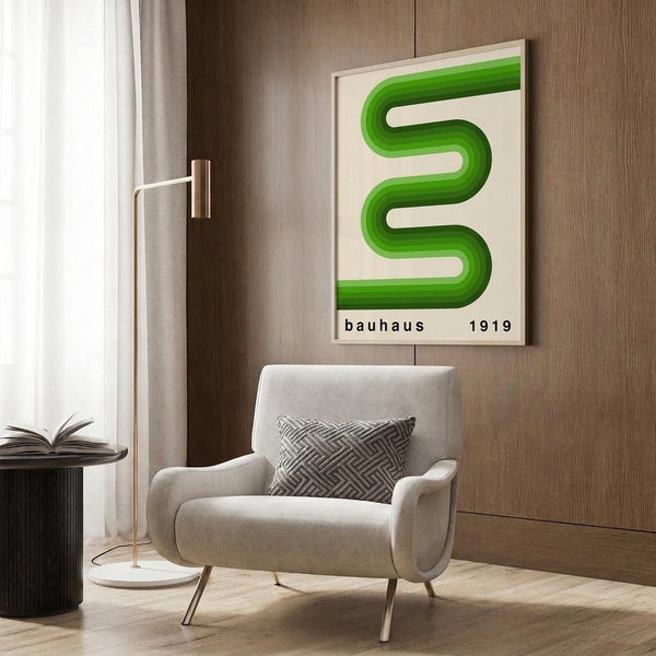 May include: Modern living room scene with beige armchair, brass floor lamp, and dark wood wall panels. Framed poster displays flowing green curved shape resembling the letter S on cream background with "bauhaus 1919" text below. Round black side table with open book and geometric patterned throw pillow complete the minimalist interior setting.