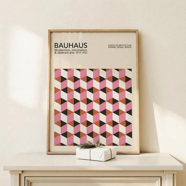 May include: Framed Bauhaus poster with geometric cube pattern in pink, orange, black and cream colors, displayed on light wood surface next to small white wrapped gift with greenery. Modern minimalist room setting with neutral walls and natural lighting.