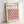 May include: Framed poster with geometric cube pattern in pink, orange, black and cream colors, featuring "BAUHAUS" text and "Modernism, minimalism & abstract arts 1919-1933" with "School of Architecture Weimar, Dessau, Berlin" in upper corner. Light wood frame leaning against white wall, showing three-dimensional isometric cube design typical of Bauhaus style.