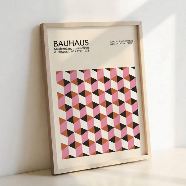 May include: Framed poster with geometric cube pattern in pink, orange, black and cream colors, featuring "BAUHAUS" text and "Modernism, minimalism & abstract arts 1919-1933" with "School of Architecture Weimar, Dessau, Berlin" in upper corner. Light wood frame leaning against white wall, showing three-dimensional isometric cube design typical of Bauhaus style.