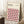May include: Framed Bauhaus-style geometric poster with pink, orange, black and white cube pattern leaning against white wall corner. Wooden frame, cream background with modernist typography. Small wooden side table with white coffee cup nearby, beige textured rug and throw blanket visible in cozy room setting.