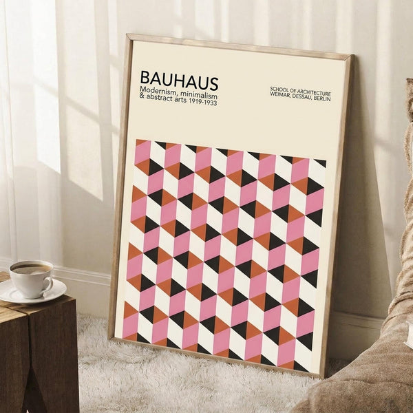 May include: Framed Bauhaus-style geometric poster with pink, orange, black and white cube pattern leaning against white wall corner. Wooden frame, cream background with modernist typography. Small wooden side table with white coffee cup nearby, beige textured rug and throw blanket visible in cozy room setting.