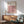 May include: Framed Bauhaus poster with pink, white, black and brown geometric cube pattern hanging above wooden sideboard. Mid-century modern living room setting with mint green upholstered armchair, small table lamp, and natural sunlight creating shadows on white wall.