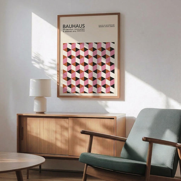 May include: Framed Bauhaus poster with pink, white, black and brown geometric cube pattern hanging above wooden sideboard. Mid-century modern living room setting with mint green upholstered armchair, small table lamp, and natural sunlight creating shadows on white wall.