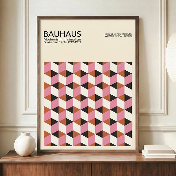 May include: Framed Bauhaus-style poster with geometric cube pattern in pink, orange, black and cream colors displayed on wooden sideboard. Modern minimalist artwork in brown frame alongside white ceramic vase and stacked books in neutral room setting with white paneled walls.