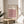 May include: Framed Bauhaus poster with geometric cube pattern in pink, coral, black, and cream colors displayed on round wooden side table next to beige upholstered chair. Natural sunlight creates shadows on neutral wall behind. Green plant leaves visible in corner. Modern minimalist interior styling with warm wood tones and soft textiles.
