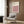 May include: Framed Bauhaus poster with pink, white, and black geometric cube pattern hanging on wood-paneled wall above beige upholstered armchair with patterned throw pillow. Modern brass floor lamp with white base stands nearby next to dark round side table with open book. Neutral tones throughout minimalist interior setting with natural lighting from window.