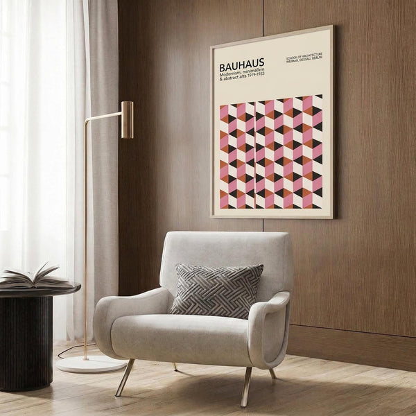 May include: Framed Bauhaus poster with pink, white, and black geometric cube pattern hanging on wood-paneled wall above beige upholstered armchair with patterned throw pillow. Modern brass floor lamp with white base stands nearby next to dark round side table with open book. Neutral tones throughout minimalist interior setting with natural lighting from window.