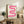 May include: Framed pink ribbon-shaped abstract poster with gradient wavy lines and "bauhaus 1919" text, displayed on rustic wooden sideboard alongside fern plant, books, ceramic vases, and brown leather armchair in warm neutral living room setting
