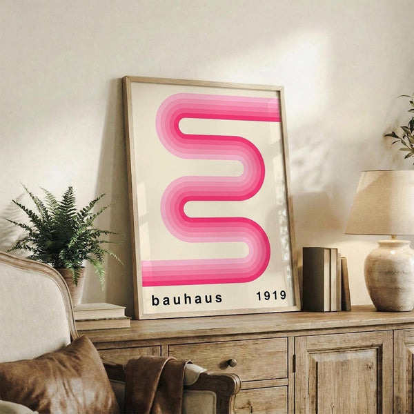 May include: Framed pink ribbon-shaped abstract poster with gradient wavy lines and "bauhaus 1919" text, displayed on rustic wooden sideboard alongside fern plant, books, ceramic vases, and brown leather armchair in warm neutral living room setting
