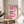 May include: Framed poster with pink gradient wavy ribbon design on cream background, displayed on round wooden side table next to beige upholstered chair. Natural sunlight creates soft shadows on white walls. Green plant leaves visible in corner. Modern minimalist interior styling with neutral tones and clean lines.