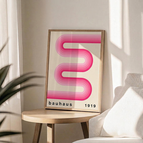 May include: Framed poster with pink gradient wavy ribbon design on cream background, displayed on round wooden side table next to beige upholstered chair. Natural sunlight creates soft shadows on white walls. Green plant leaves visible in corner. Modern minimalist interior styling with neutral tones and clean lines.