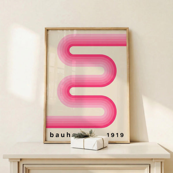 May include: Framed poster with pink gradient ribbon design in wavy S-curve pattern against cream background, displayed on light wood console table next to small wrapped gift with greenery sprig. Thin natural wood frame with glass front, soft shadows on neutral wall behind.