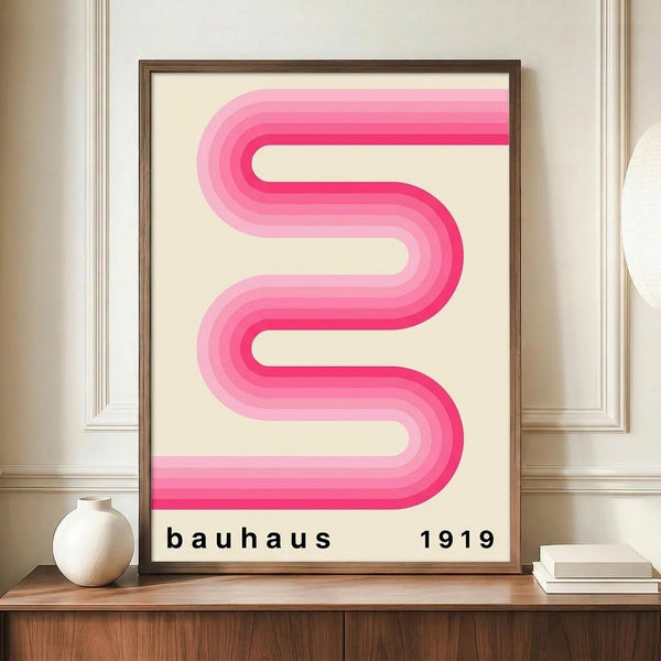 May include: A framed poster featuring a flowing pink ribbon design with gradient stripes creating an S-shaped curve on cream background, with "bauhaus 1919" text below. The brown wooden frame sits on a wooden sideboard next to a white ceramic vase and books, against beige paneled walls in an elegant interior setting.