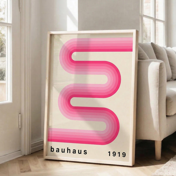 May include: Framed poster with curved pink gradient ribbon design on cream background, text reading "bauhaus 1919" below. Light wood frame leaning against white wall next to beige upholstered chair with throw pillows. Bright natural light from tall windows creates soft shadows on hardwood floor.
