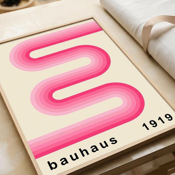 May include: Pink wavy ribbon design poster with cream background displayed on wooden surface next to rolled white fabric. The minimalist artwork features flowing curved lines in gradient pink tones from light to deep magenta, with "bauhaus 1919" text at bottom in black letters. The print appears in a thin wooden frame against neutral textured materials.