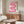 May include: Pink gradient ribbon-shaped design forming flowing S-curve on cream background in thin brown frame, displayed above wood sideboard with sliding doors. Modern white sculptural chair and glass coffee table in foreground. Bauhaus 1919 text at bottom. Minimalist room with beige walls, natural lighting creating soft shadows on polished floor.