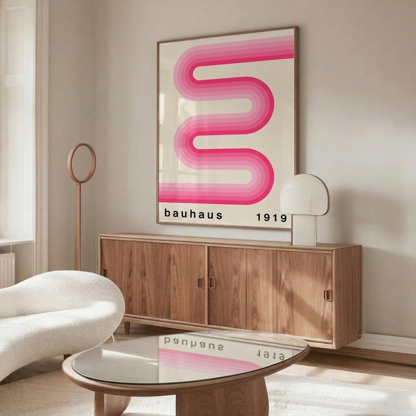 May include: Pink gradient ribbon-shaped design forming flowing S-curve on cream background in thin brown frame, displayed above wood sideboard with sliding doors. Modern white sculptural chair and glass coffee table in foreground. Bauhaus 1919 text at bottom. Minimalist room with beige walls, natural lighting creating soft shadows on polished floor.